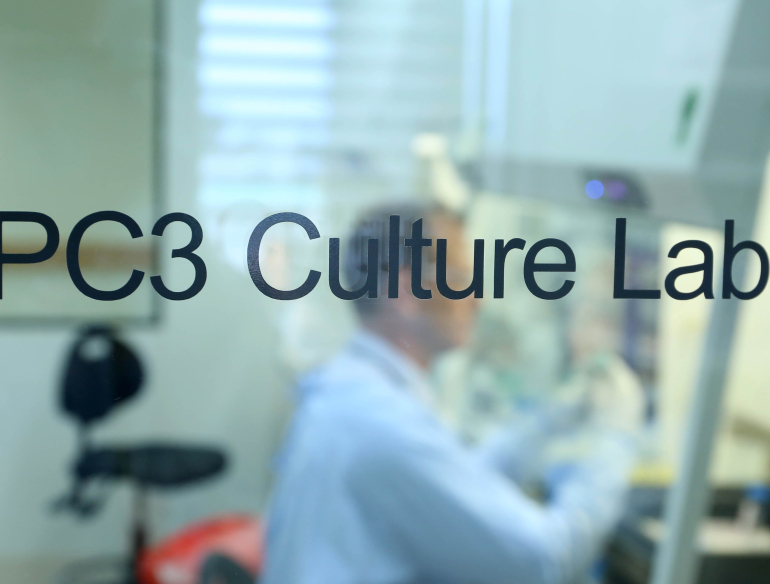 Close up of a sign door "PC3 Culture Lab". Person in the background, working at a hood. 