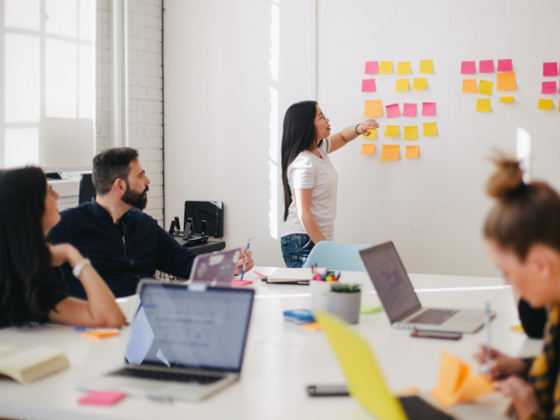 Photo of group of people around a table having a meeting, while one person is in the front pointing at post it notes on the wall. Credit: Unsplash