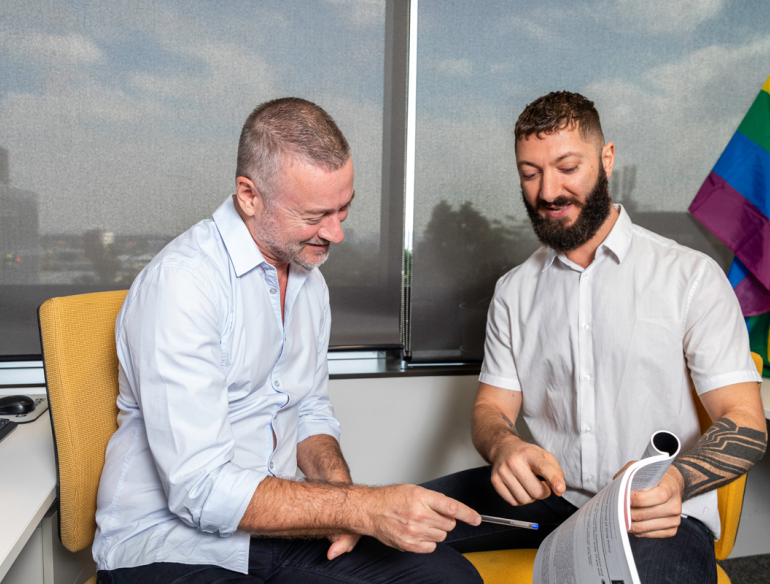 Two men sitting and reading a magazine together, rainbow pride flag in background