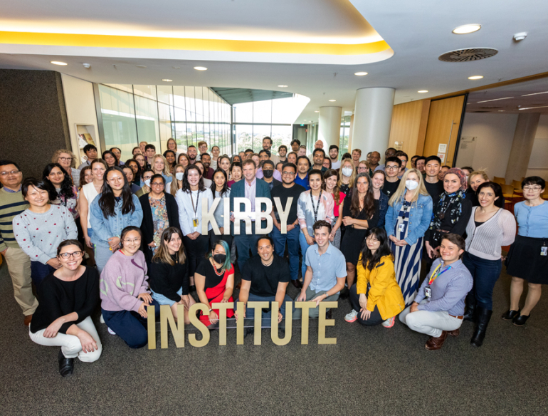 Kirby Institute staff posing with "Kirby Institute" sign. Kirby researcher presenting to colleagues. Credit: Bec Lewis/Kirby Institute.
