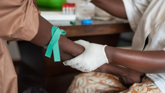 Two dark skinned people at a medical examination, where one is taking a blood sample from the other. Credit: Shutterstock.