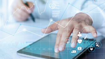 Graphs and medical symbols overlayed over a medical staff member writing, one hand touching a tablet. Credit: AdobeStock.