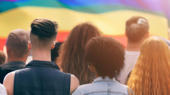 Group of diverse LGBTQ crowd from behind, in front of a rainbow flag.
