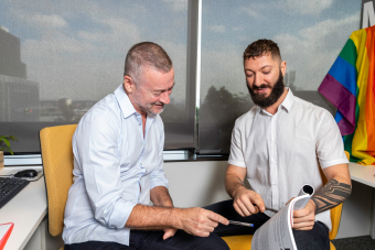 Two men sitting and reading a magazine together, rainbow pride flag in background