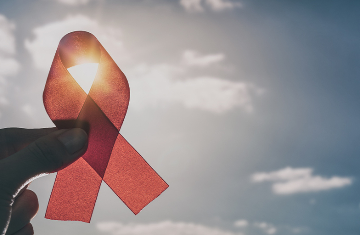 An HIV red ribbon is held up close to the camera in front of a blue, sunlit sky